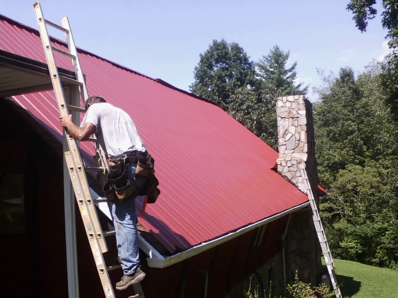 Roofer on a ladder beside a red metal roof and stone chimney — Soffit & Fascia Repair by Roofing Brothers in Zapata